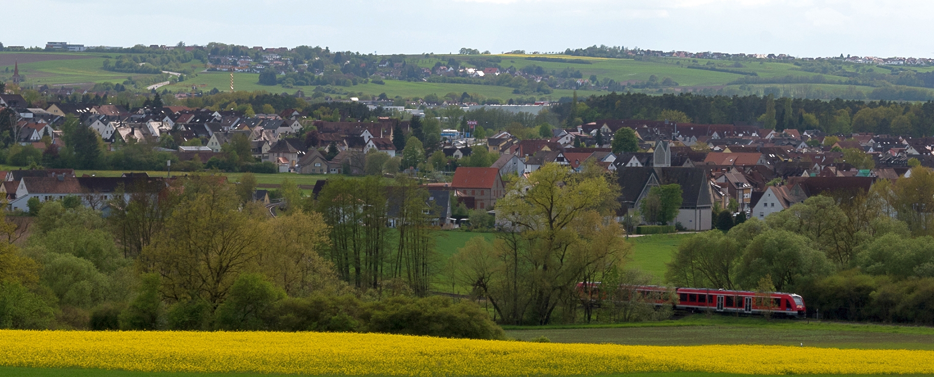 Blick auf die Ortschaft Forth mit Rapsfeldern im Vordergrund, der Gräfenbergbahn zwischen Bäumen und dem hellblauen Himmel im Hintergrund